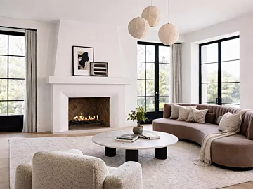 EcoSmart Fire Heritage 50 Grate installed in a white built-in fireplace with visible flame, set in a bright contemporary living room featuring a curved sofa, marble coffee table and large black-framed windows.